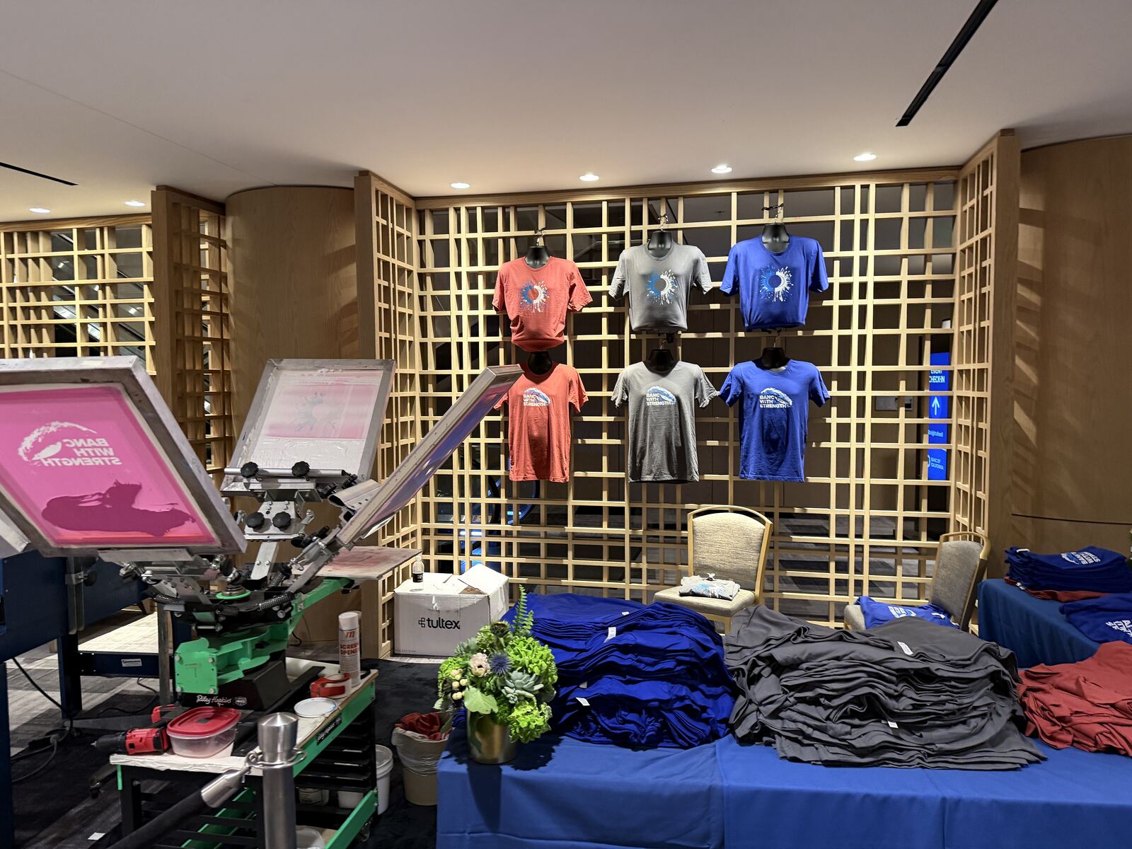 Professional print booth showcase with hanging apparel on a gridded display wall and screen printing equipment with sample products